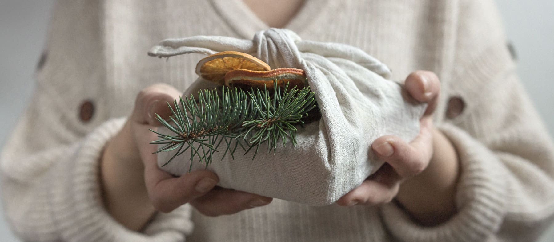 Person holding a gift wrapped in white cloth with a pine tree branch